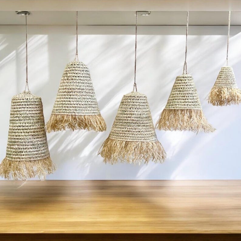 May include: Five natural woven pendant lights with fringe trim hang from the ceiling against a white wall. The lights are different sizes, with the largest light at the top and the smallest light at the bottom.