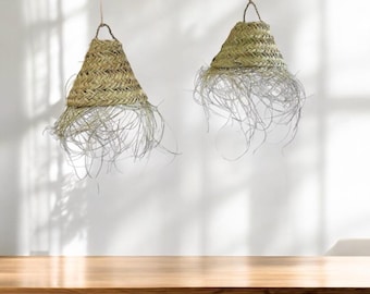 May include: Two woven straw pendant lights with a fringe of natural fibers hanging from a white wall. The lights are hanging over a wooden surface.