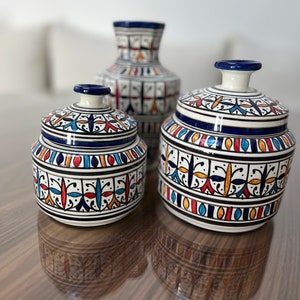 Puede incluir: Tres recipientes de cerámica con intrincados patrones geométricos en azul, naranja y rojo. Los recipientes son blancos con un acabado brillante. El recipiente más grande es un jarrón, y los dos recipientes más pequeños tienen tapas.