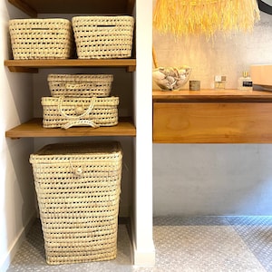 Puede incluir: Un conjunto de cuatro cestas de almacenamiento tejidas en color beige natural. Las cestas están apiladas en un estante de madera. Las cestas tienen un patrón tejido y están hechas de materiales naturales.