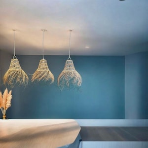 May include: Three woven pendant lights with a natural, light brown color hang from the ceiling against a blue wall. The lights are in a row and are evenly spaced.