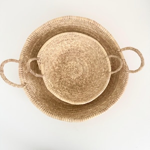 Pode incluir: Duas cestas redondas de palha tecida. A cesta maior tem cerca de 46 cm de diâmetro e apresenta duas alças curvas. A cesta menor, com cerca de 30 cm de diâmetro, está dentro da maior, também com alças. Ambas são castanho claro.