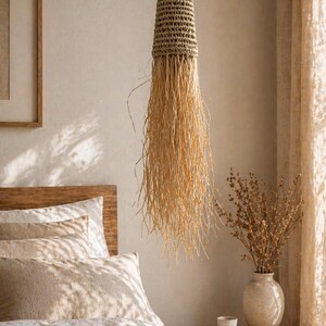 Op de afbeelding: Een hangende geweven pendel met een cilindrische bovenkant en lange, vloeiende strengen natuurlijke vezels. De lamp is opgehangen aan een dun koord. Het kleurenpalet is neutraal, met beige en bruine tinten, wat een warme en uitnodigende sfeer creëert.