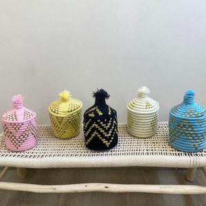 Op de afbeelding: Vijf decoratieve geweven manden met deksels in verschillende kleuren, waaronder roze, geel, zwart, wit en blauw. De manden zijn gerangschikt op een licht houten bankje met een geweven zitting.