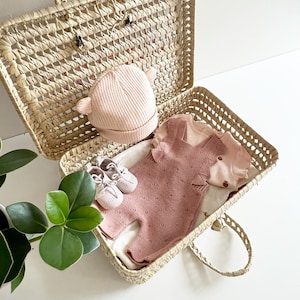 Puede incluir: Una cesta de mimbre tejida con asa, abierta para revelar un conjunto de bebé de punto rosa, un gorro de punto rosa y un par de zapatos de bebé blancos.