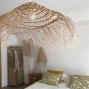 May include: A woven, natural fiber pendant light fixture with a wide, fringed shade. The light hangs from a white cord, casting a warm glow. The bedroom setting includes a bed with floral pillows and a wooden nightstand.