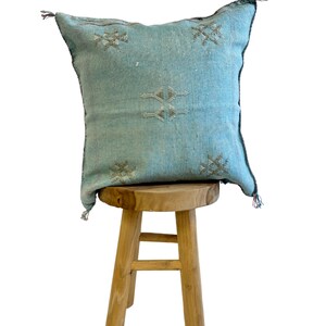 May include: A light blue throw pillow with a geometric pattern sits on a wooden stool. The pillow has a fringe border and is made of a soft, woven fabric.