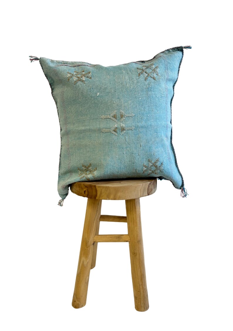 May include: A light blue throw pillow with a geometric pattern sits on a wooden stool. The pillow has a fringe border and is made of a soft, woven fabric.