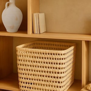 Puede incluir: Una cesta de almacenamiento tejida hecha de materiales naturales. La cesta es de color marrón claro y tiene una forma cuadrada. Está sentada en un estante de madera.