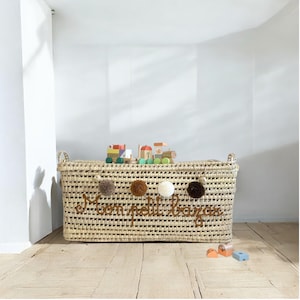 May include: A rectangular woven basket with handles, holding wooden toy train and blocks. The basket has the text "mon petit bazar" and is decorated with pom-poms. The basket is on a wooden floor.