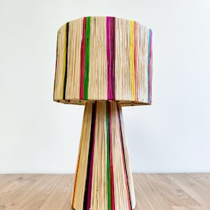 Puede incluir: Una lámpara de mesa colorida con una pantalla y una base tejidas. La pantalla está hecha de fibras naturales y tiene rayas de color rosa, rojo, amarillo, verde y negro. La base tiene forma de cono y también está tejida con los mismos colores.