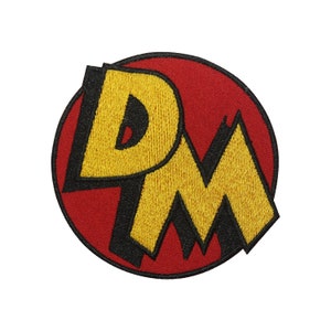 May include: An embroidered patch featuring the letters "D.M." in a stylised font, with a red background and yellow lettering.