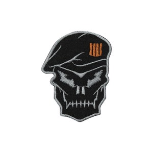 May include: Embroidered patch featuring a stylized skull wearing a black beret. The skull has detailed silver stitching for the facial features and a row of orange bars on the beret. The patch is designed with a black background.