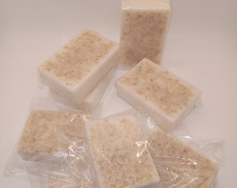 May include: A group of six white soap bars wrapped in clear plastic. The soap bars have a light brown, grainy texture and some have a honeycomb design.