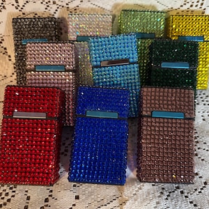 May include: A collection of bedazzled cigarette cases in various colors, including red, blue, brown, and green. Each case is covered in small, sparkling rhinestones and features a silver-tone clasp.