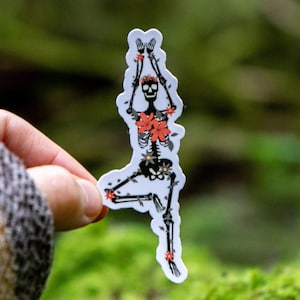May include: A sticker of a skeleton doing yoga with flowers on its body. The sticker is white with black and red details. The sticker is being held by a hand in a forest setting.