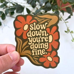 May include: A sticker with a retro floral design in shades of orange, brown, and olive green. The sticker features the text "slow down, you're doing fine" in a cream-coloured font, perfect for adding a touch of positivity to any item.