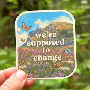 Puede incluir: Una pegatina cuadrada con esquinas redondeadas que presenta una ilustración de un paisaje con una montaña y un prado de flores silvestres bajo un cielo azul. El texto "we're supposed to change" está impreso en blanco en el centro, junto a mariposas naranjas y azules.