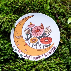 May include: A white sticker featuring a yellow crescent moon with a sleeping face and a cluster of pink, red, and yellow flowers. The text "Take a moment to breathe." is printed on the sticker.