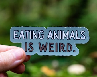 Puede incluir: Una pegatina azul con texto blanco que dice "Comer animales es raro." La pegatina está siendo sostenida por una mano en un entorno de bosque.