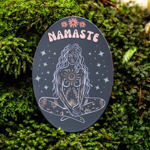 May include: A black oval sticker with a gold outline and a white illustration of a woman sitting in a yoga pose. The woman has long hair and is surrounded by stars and other celestial symbols. The sticker says "NAMASTE" in pink letters.
