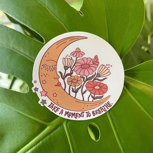 Peut inclure: Un autocollant avec un croissant de lune avec un visage et des fleurs. La lune est orange et les fleurs sont roses, rouges et jaunes. Le texte "Take a moment to breathe." est &eacute;crit sous la lune.