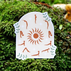 May include: A sticker with a white background and a brown sun in the center. The sun has rays that extend outward. Around the sun are 12 brown silhouettes of people in different yoga poses. The sticker has a rounded arch shape at the top and a green leafy design at the bottom.