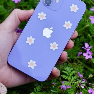 May include: A purple iPhone with a white apple logo and white daisy stickers on the back. The phone is being held in a hand against a background of purple flowers.