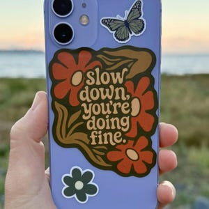 May include: A lilac phone case with a floral design and the text "slow down, you're doing fine." The case also features a butterfly and a small green flower sticker. The phone case is held in a person's hand.