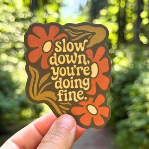 May include: A die-cut sticker with a dark brown background featuring orange flowers and yellow centers. The text in the center reads, "slow down, you're doing fine." The sticker has a wavy, organic border shape and a matte finish with a textured appearance.