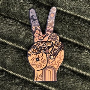 May include: A die-cut sticker shaped like a hand making a peace sign. The design features a warm color palette with illustrations of a butterfly, a crescent moon, stars, a sun, flowers, an eye, a rainbow, and trees. The word Peace is written.