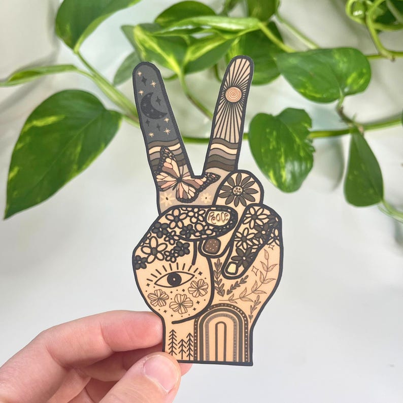 May include: A die-cut sticker shaped like a hand making a peace sign, featuring intricate line art in shades of beige, brown, and black. The design includes a crescent moon, sun, butterfly, flowers, an eye, and the word peace written on the palm.