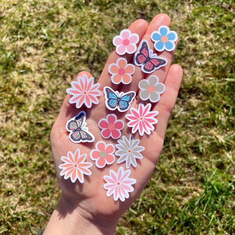 Op de afbeelding: Een verzameling kleurrijke bloemen- en vlinderstickers. De stickers zijn roze, blauw en bruin. Ze zijn in een hand gerangschikt, met de bloemen naar boven en de vlinders naar beneden.