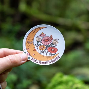 Peut inclure: Un autocollant blanc avec une lune en croissant jaune avec un visage endormi et des fleurs devant. Le texte "Take a moment to breathe" est &eacute;crit sous la lune.