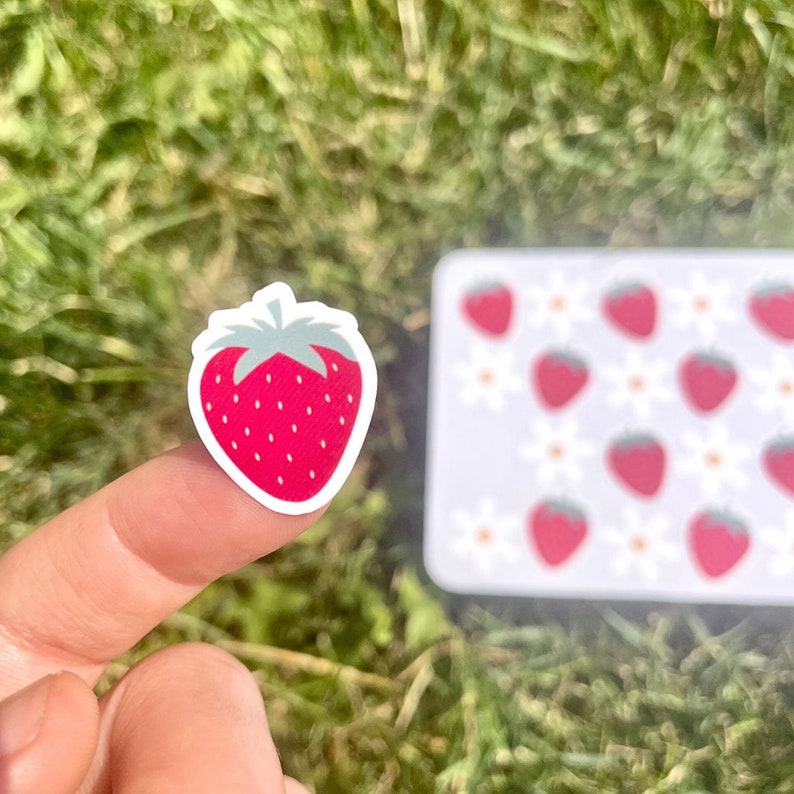 May include: A red strawberry sticker with a green stem and leaves. The sticker is on a finger against a green grassy background. There is a sheet of stickers in the background with a pattern of strawberries and white flowers.