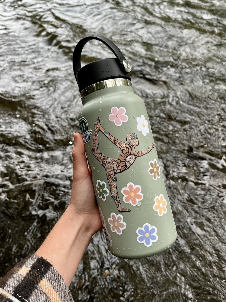 May include: A green reusable water bottle with a black lid and handle. The bottle is decorated with colorful flower and floral pattern stickers. One sticker features a woman doing a yoga pose.