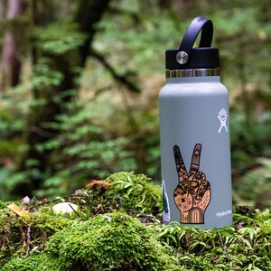May include: A light gray Hydro Flask water bottle with a black lid and handle. The bottle features a white logo and a detailed sticker of a hand making a peace sign, decorated with intricate patterns including flowers, suns, and geometric shapes.