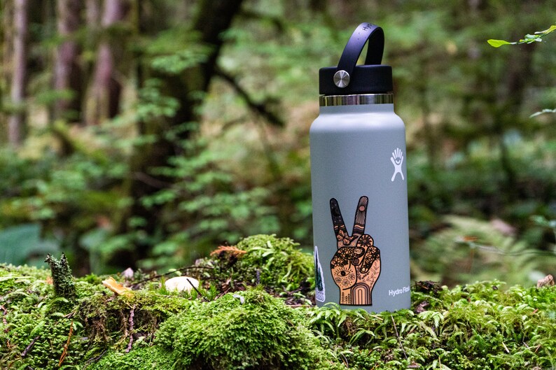 May include: A light gray Hydro Flask water bottle with a black lid and handle. The bottle features a white logo and a detailed sticker of a hand making a peace sign, decorated with intricate patterns including flowers, suns, and geometric shapes.