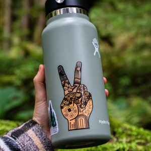 May include: A sage green Hydro Flask water bottle with a black lid and a large sticker of a hand making a peace sign. The sticker features intricate line art of flowers, a sun, a moon, and an eye. A smaller sticker of pine trees is visible.