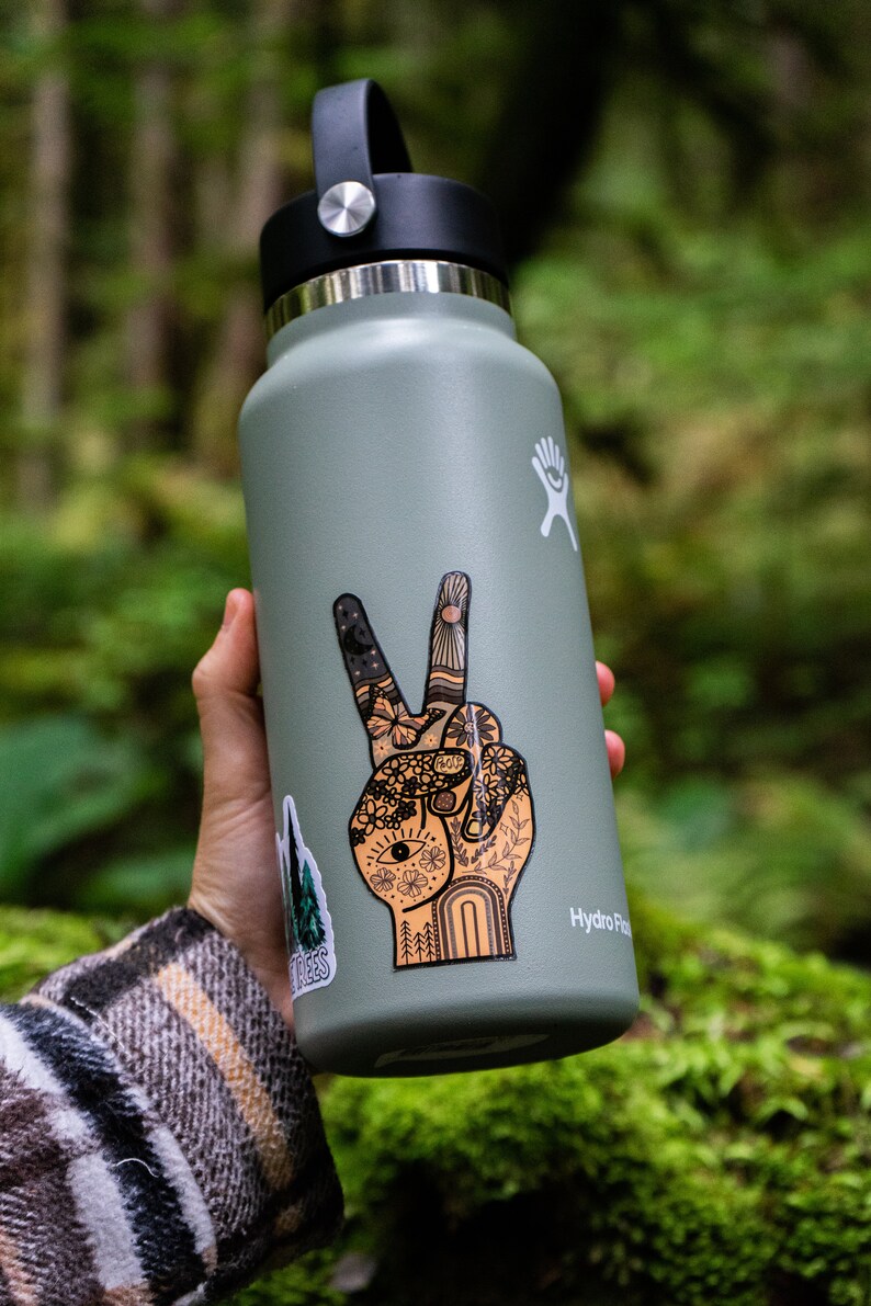 May include: A sage green Hydro Flask water bottle with a black lid and a large sticker of a hand making a peace sign. The sticker features intricate line art of flowers, a sun, a moon, and an eye. A smaller sticker of pine trees is visible.