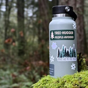 May include: A light green water bottle with a black lid and handle. The bottle has stickers that say "TREE-HUGGER PEOPLE-AVOIDER" and "TAKE ME TO THE TREES" with tree and flower graphics. The bottle is sitting on moss.