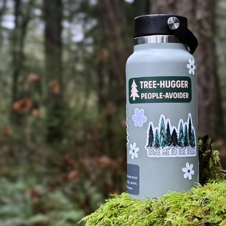 May include: A light green water bottle with a black lid and handle. The bottle has stickers that say "TREE-HUGGER PEOPLE-AVOIDER" and "TAKE ME TO THE TREES" with tree and flower graphics. The bottle is sitting on moss.