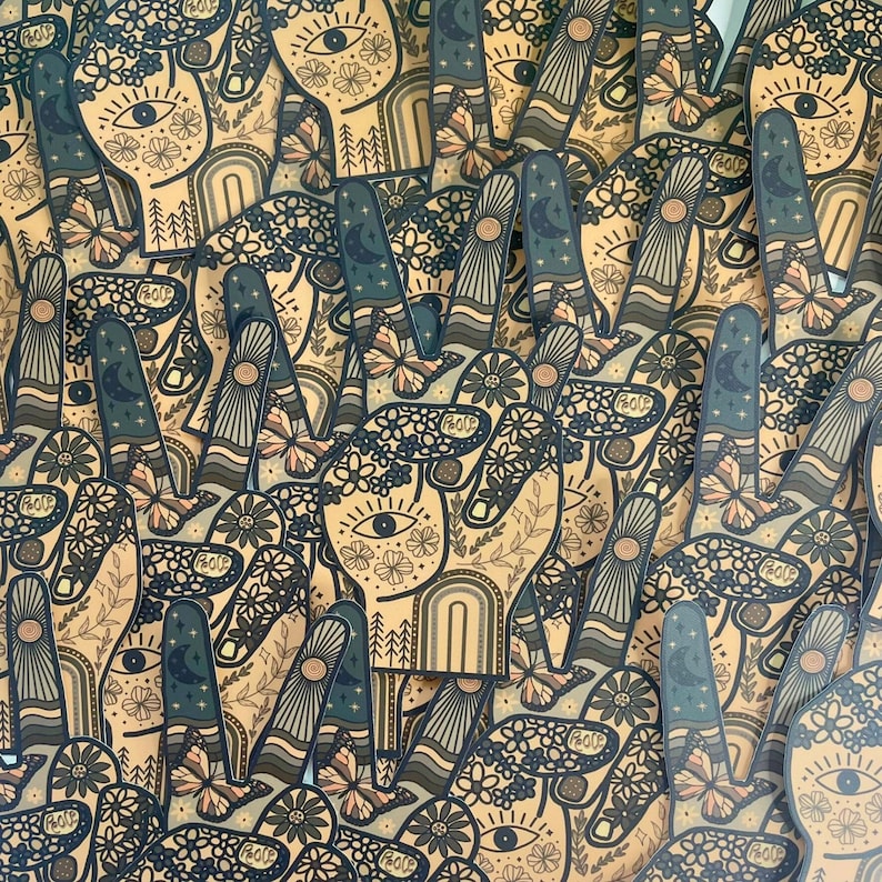 May include: A collection of die-cut stickers shaped like a hand making a peace sign. Each sticker features a tan background with dark blue line art depicting flowers, a central eye, a crescent moon, butterflies, and the word PEACE on the thumb.