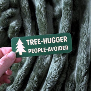 May include: A green rectangular sticker with rounded corners. The sticker features a white tree graphic and the text "TREE-HUGGER PEOPLE-AVOIDER" in white. The sticker is held against a green, textured background.