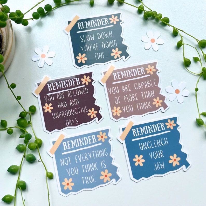 May include: Five colourful reminder stickers with a daisy design and encouraging messages. The messages are: "Slow down, you're doing fine", "You are allowed bad and unproductive days", "Not everything you think is true", "You are capable of more than you think", and "Unclench your jaw".