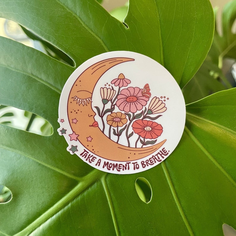 Peut inclure: Un autocollant avec un croissant de lune avec un visage et des fleurs. La lune est orange et les fleurs sont roses, rouges et jaunes. Le texte "Take a moment to breathe." est &eacute;crit sous la lune.