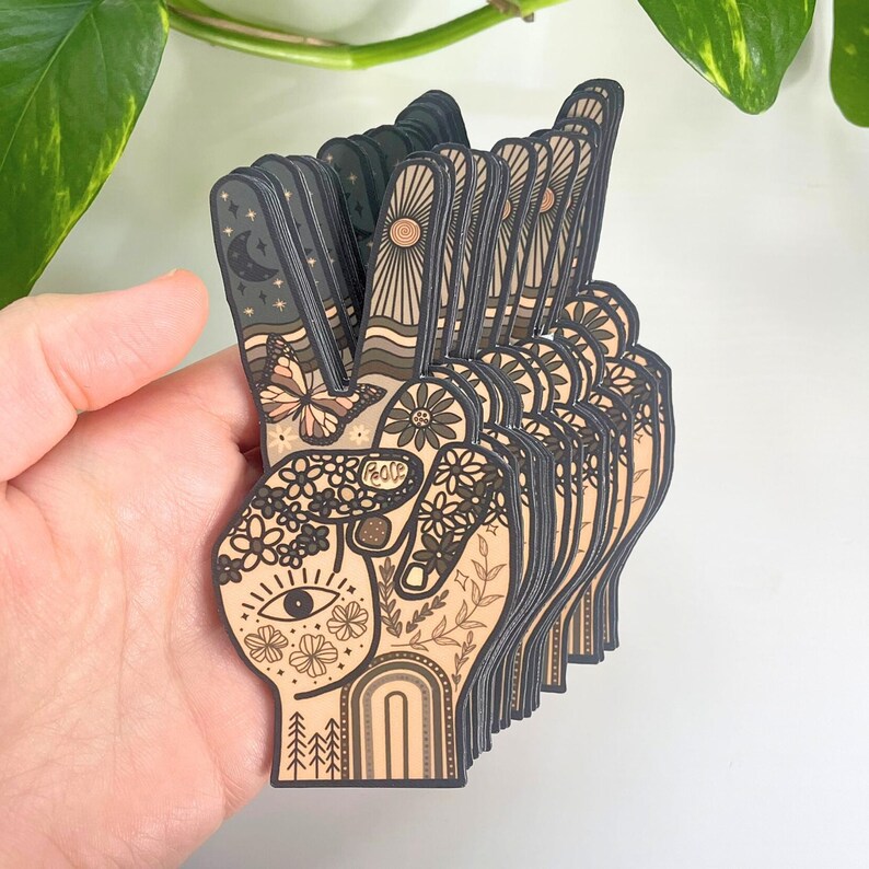 May include: A stack of die-cut stickers shaped like a hand making a peace sign. The design features a tan background with black line art including a butterfly, flowers, an eye, a crescent moon, a sun, a rainbow, and the word peace written on a finger.
