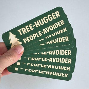 May include: A stack of green rectangular cards with rounded corners. The cards have white text that reads "TREE-HUGGER PEOPLE-AVOIDER" and a tree graphic. The cards are held in a hand.