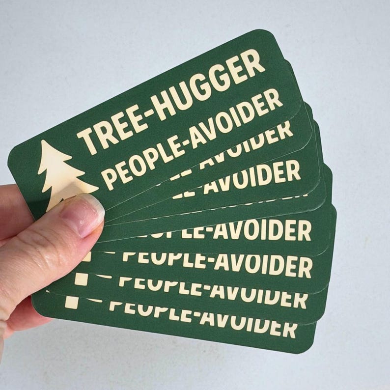 May include: A stack of green rectangular cards with rounded corners. The cards have white text that reads "TREE-HUGGER PEOPLE-AVOIDER" and a tree graphic. The cards are held in a hand.