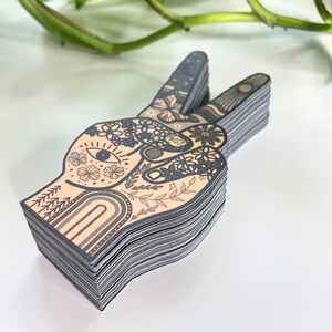 May include: A stack of die-cut stickers shaped like a hand making a peace sign. The design features a peach-colored background with black line art including flowers, an eye, a butterfly, a rainbow, and the word Peace written on a finger.